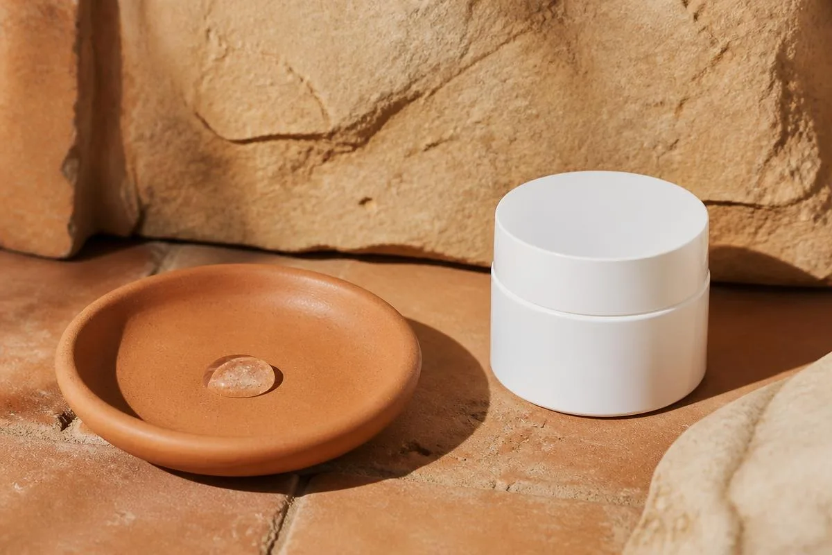 A pea-sized dose of clear gel in a terracotta dish beside a plain white cream jar on warm clay tile