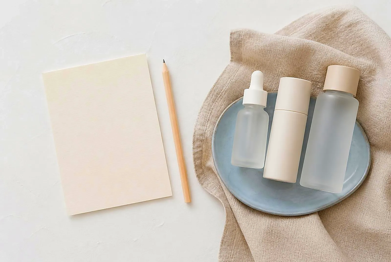A blank cream notecard and pencil beside three unlabeled bottles grouped on a matte ceramic dish over oatmeal linen