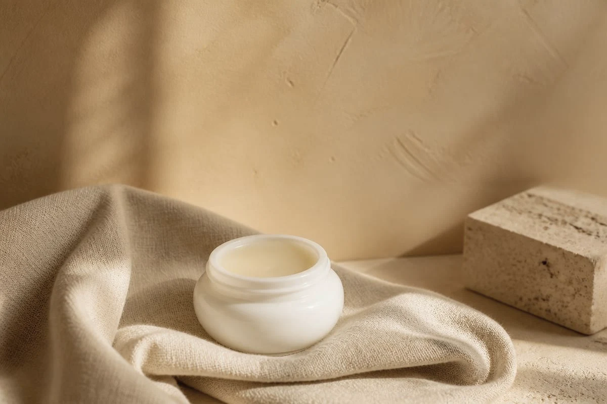 Open ceramide balm jar on folded oatmeal linen beside a travertine block against a soft honey wall, for repairing a damaged skin barrier