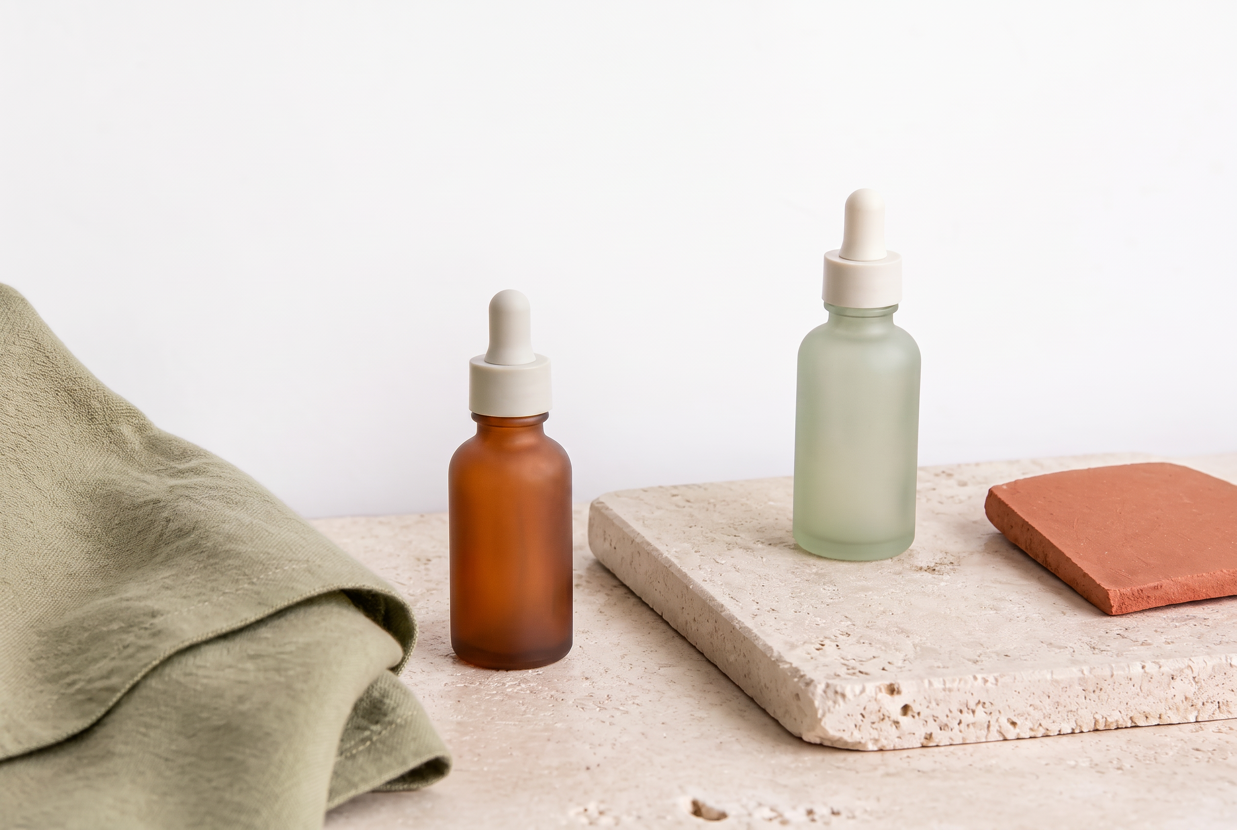 An amber dropper bottle and a frosted sage serum bottle set well apart on cream travertine beside sage linen and a terracotta tile