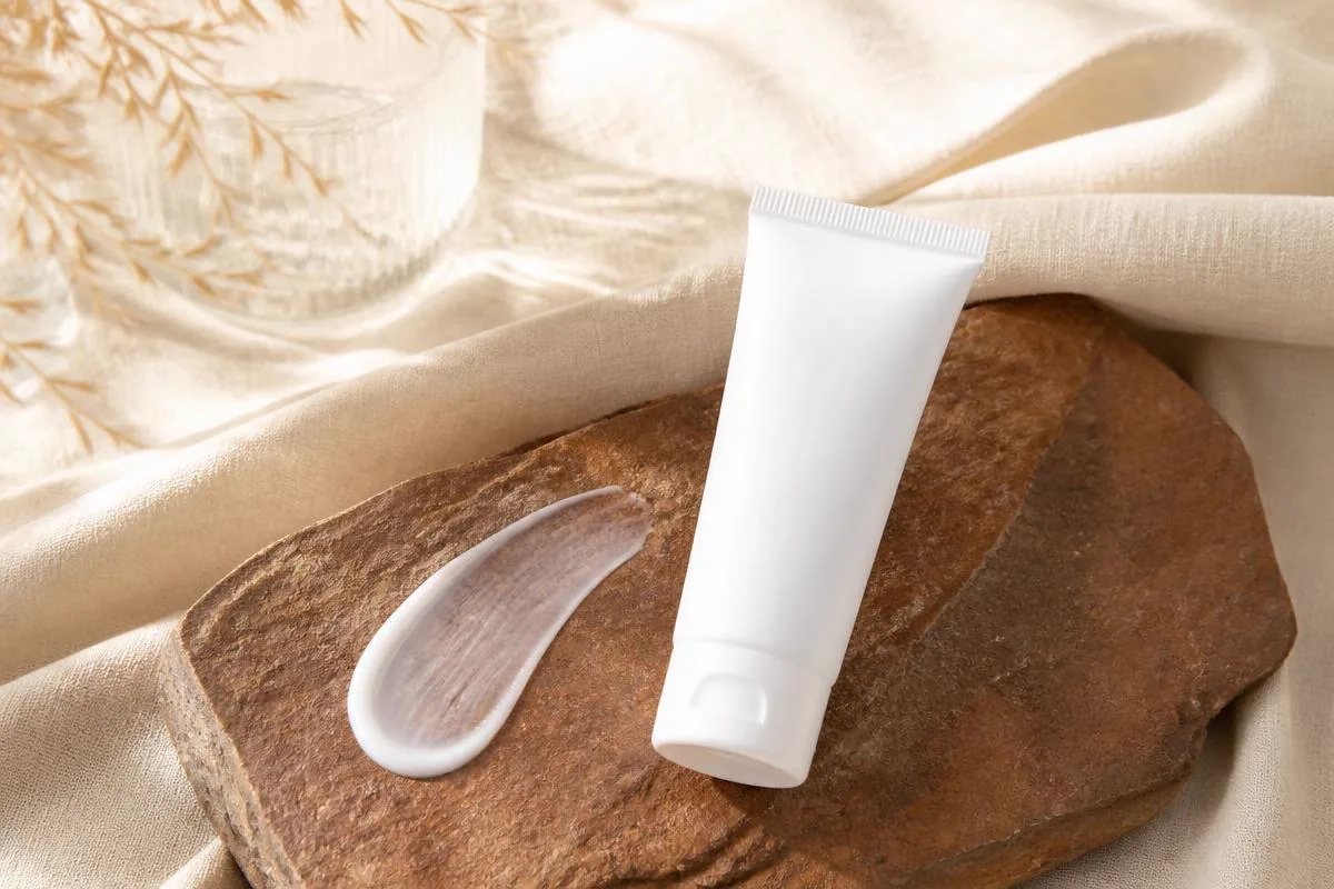 White sunscreen tube and a sheer swatch smoothed across warm deep brown stone, showing no white cast