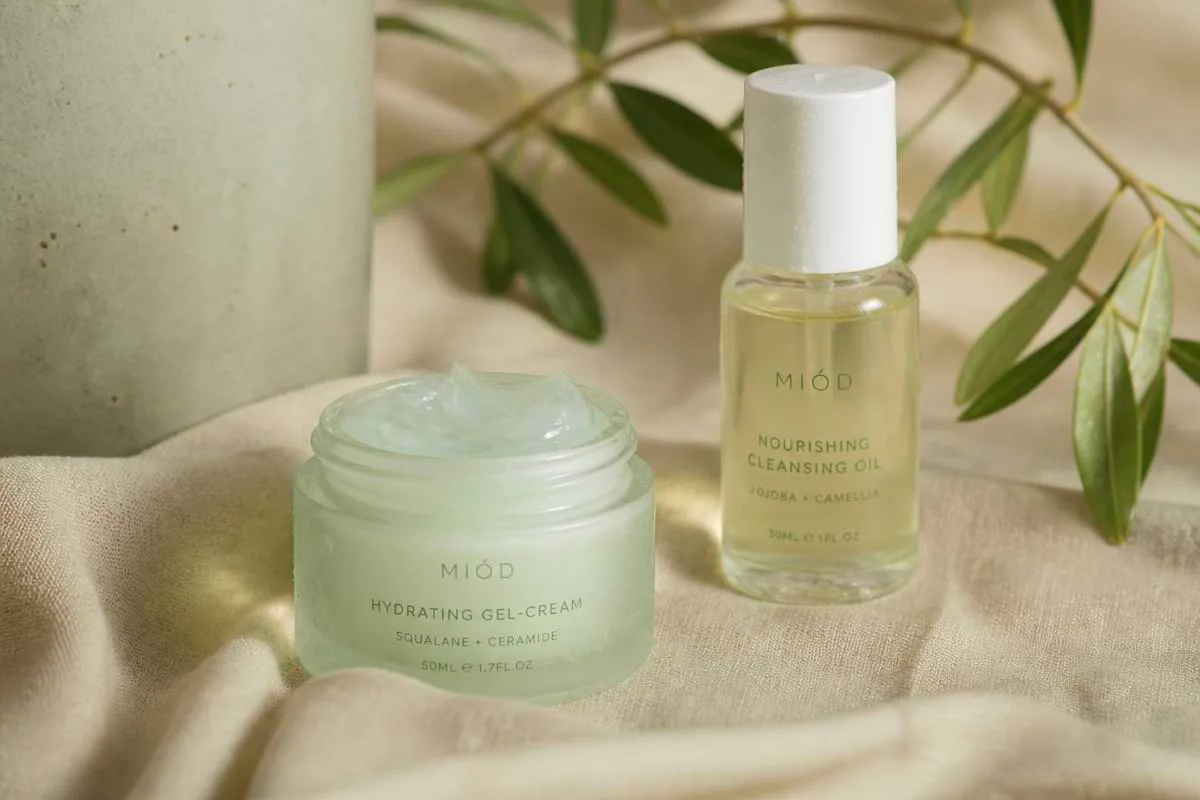 Frosted jar of hydrating gel-cream and a cleansing oil bottle on sand linen with olive stems, for a hydration-layering routine