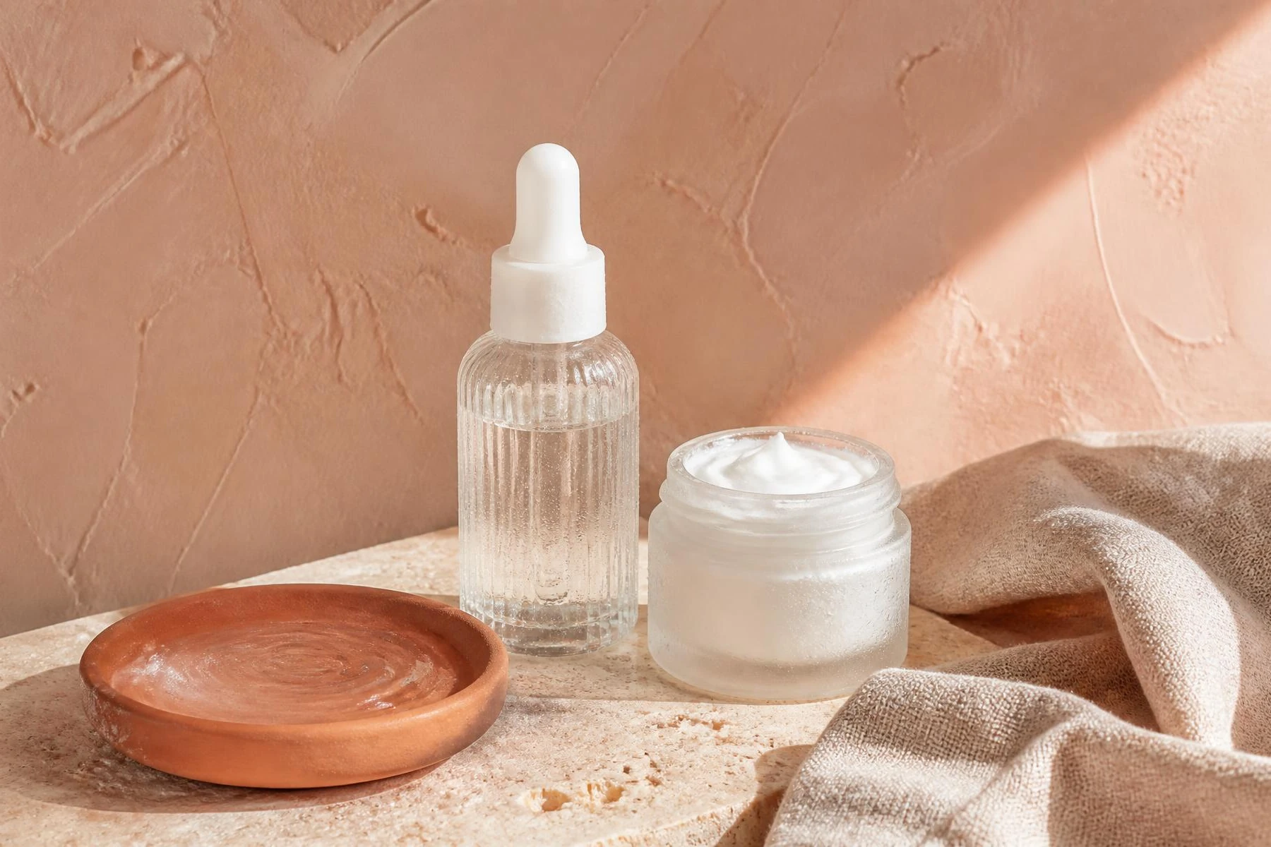 Frosted jar of hydrating gel-cream and a cleansing oil bottle on sand linen with olive stems, for a hydration-layering routine