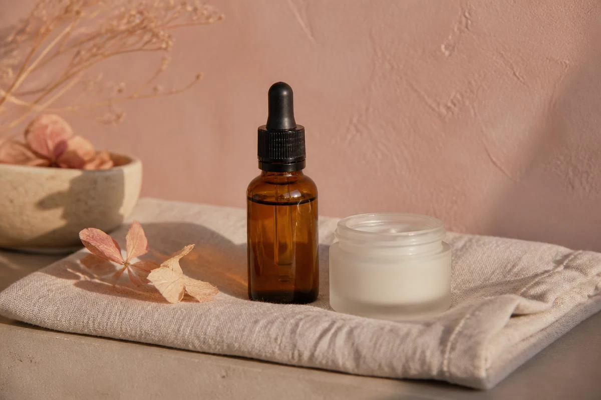 Amber vitamin C dropper and a cream tube on a warm sand ledge against a soft blush wall, for fading dark spots