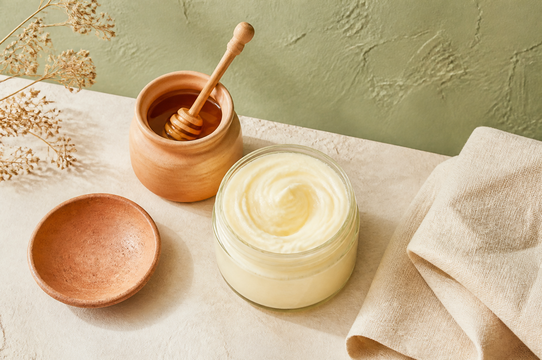 Small jars of tallow balm and a honey dipper on cream linen against warm travertine, showing how to layer tallow skincare