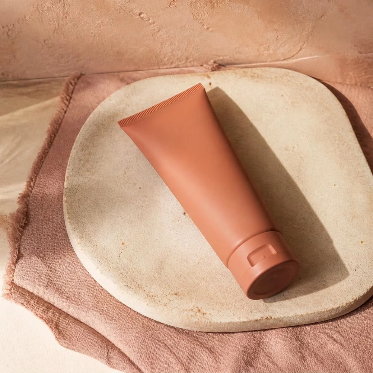 Soft clay moisturizer tube resting flat on a pale stone tray over a dusty rose linen runner against a warm apricot wall. Bottle finish: Clay.