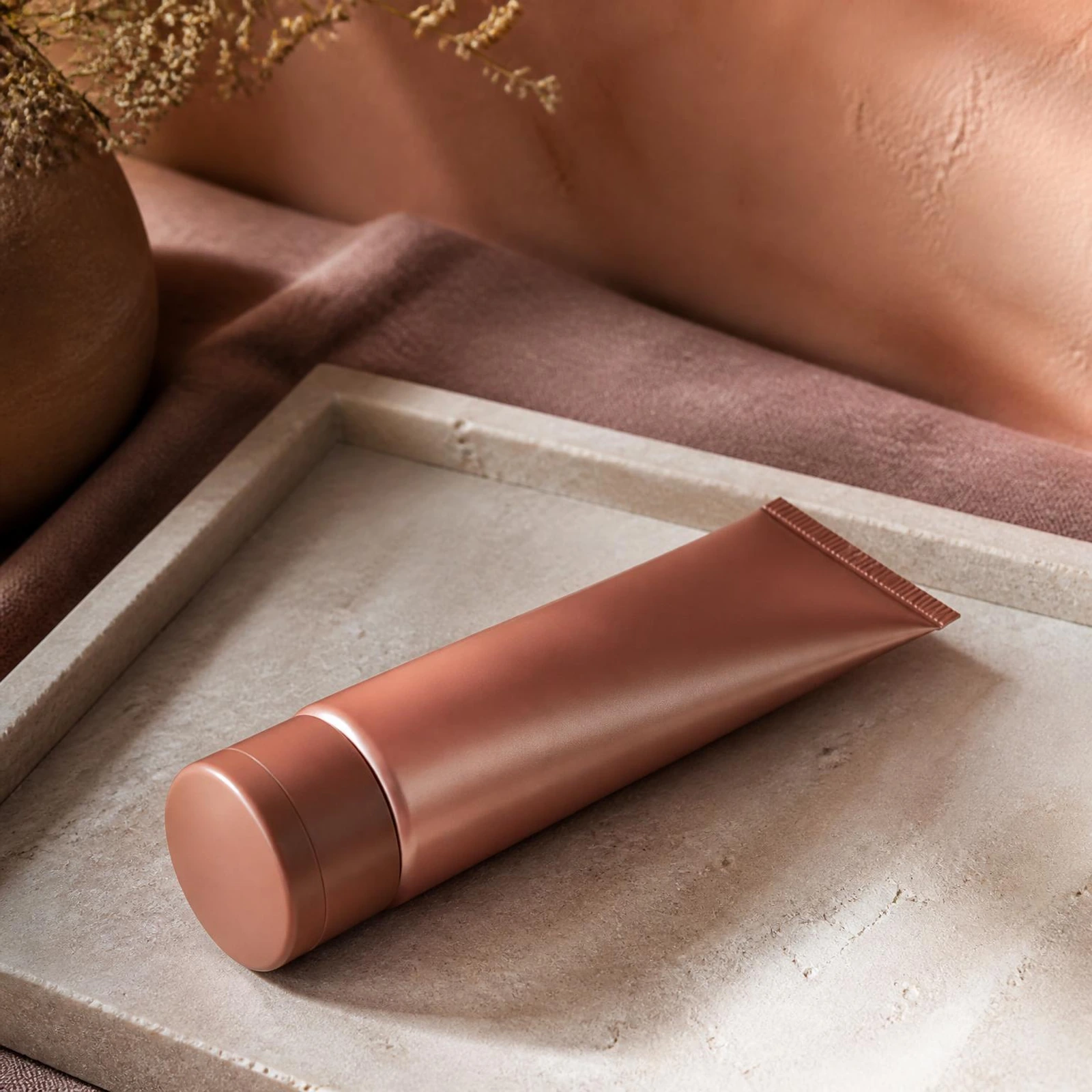 Soft clay moisturizer tube resting flat on a pale stone tray over a dusty rose linen runner against a warm apricot wall. Bottle finish: Clay.
