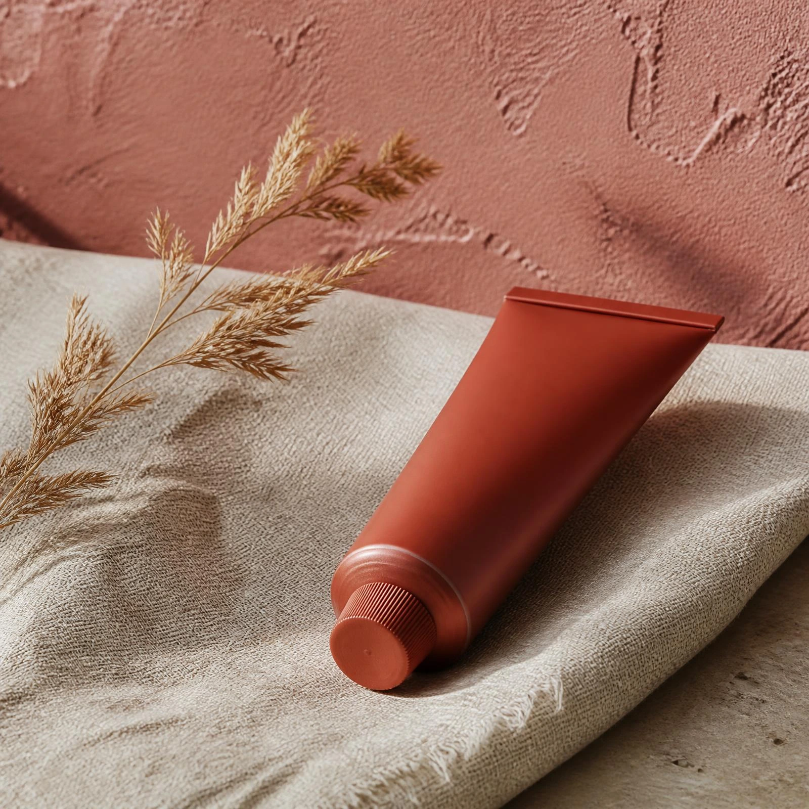 Clay-toned moisturizer tube lying on a cream linen runner with a dried grass sprig against a dusty rose wall. Bottle finish: Clay.