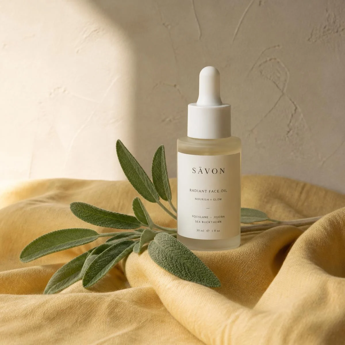 Frosted glass dropper bottle on folded butter yellow linen with a sage sprig against a warm oat plaster wall. Bottle finish: Frosted.