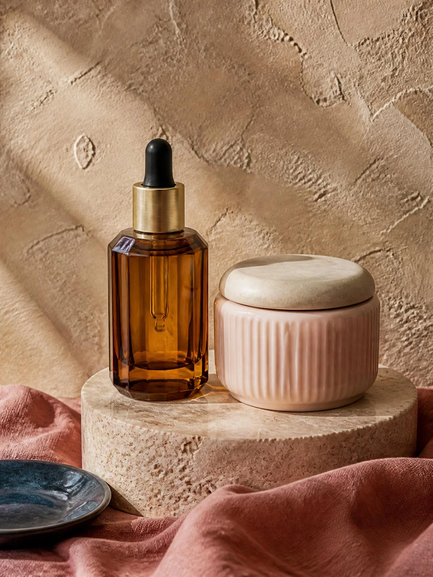 Skincare still life: amber serum dropper bottle and pale blush cream jar on a honed travertine plinth, warm sand plaster wall, dusty rose linen and a slate blue dish. Bottle finish: Amber + blush. — skincare routine quiz