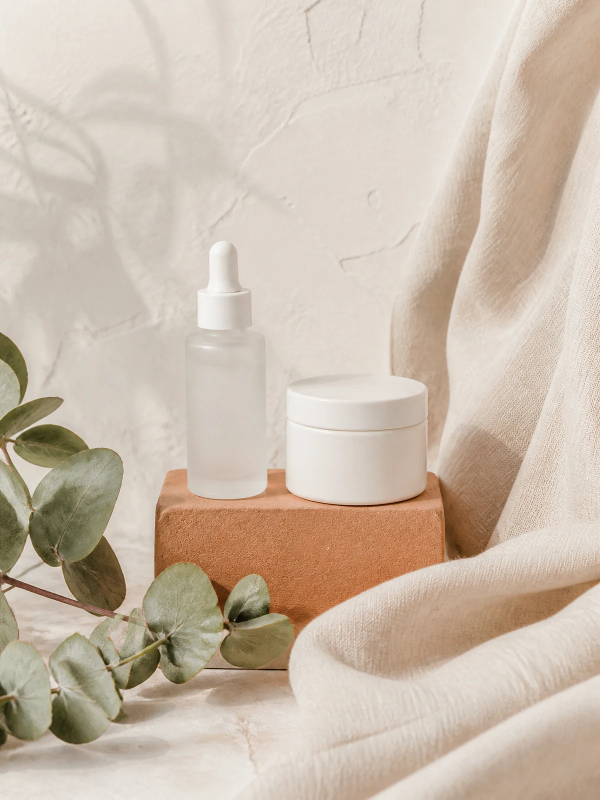 Previous hero frame: frosted serum dropper bottle and white cream jar on a clay block against a pale cream plaster wall