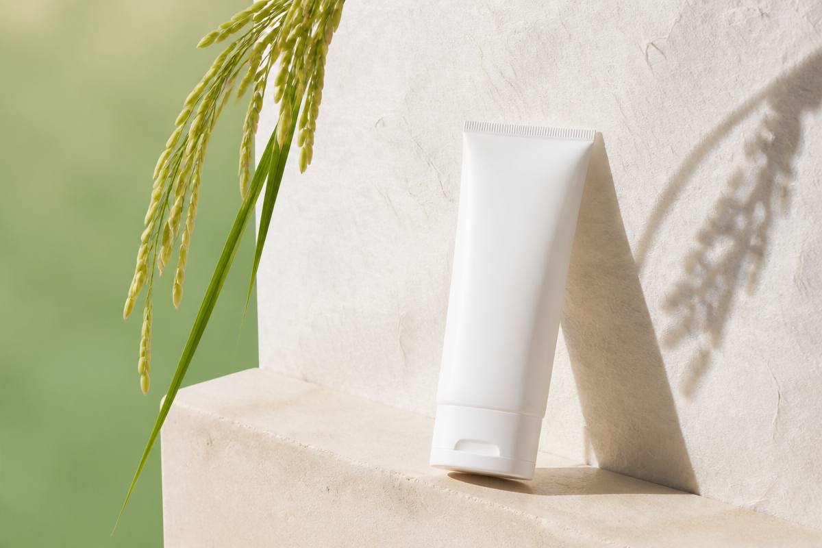 Unlabeled white sunscreen tube leaning on a pale stone block outdoors beside a green rice stalk in bright daylight