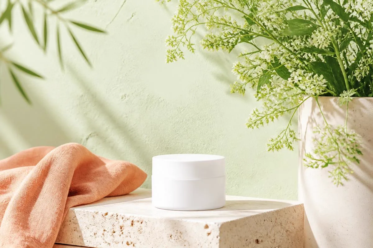Plain white cream jar with its lid off on a pale stone bathroom shelf beside a folded gray washcloth