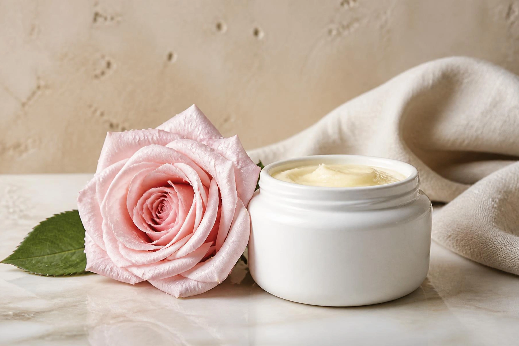 Open white ceramic jar of soothing barrier balm beside a single pale pink garden rose on cream marble with folded linen