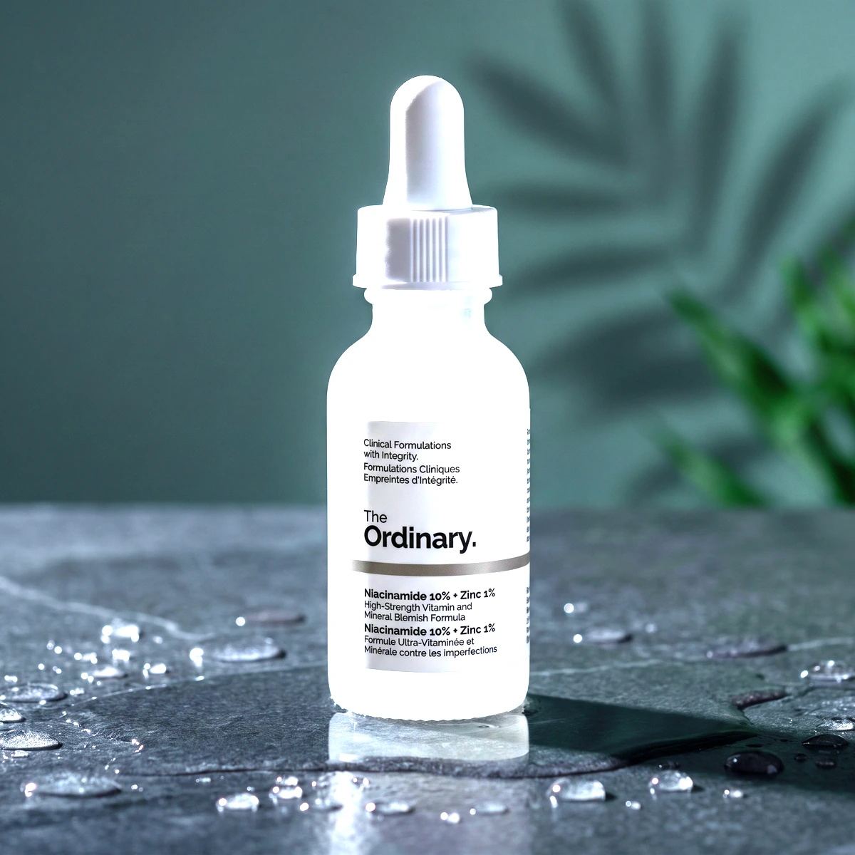 The Ordinary Niacinamide 10% + Zinc 1% dropper bottle on wet dark stone, with clear droplets and a leaf shadow