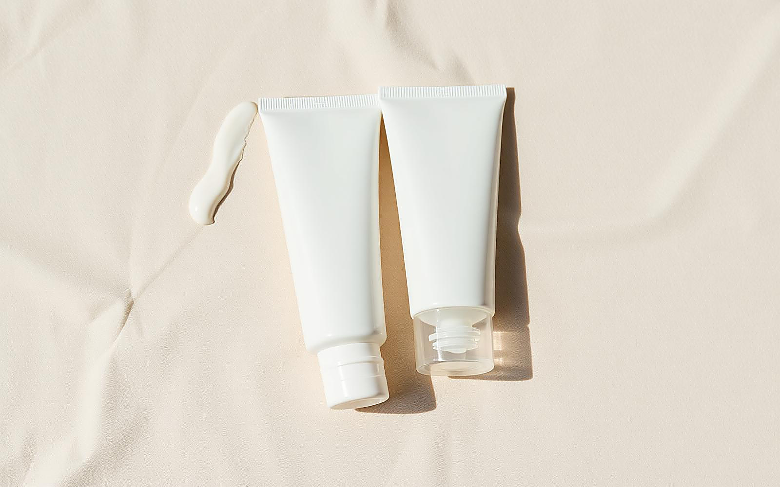 Two plain white sunscreen tubes side by side on pale sand linen with a thick cream swatch beside them