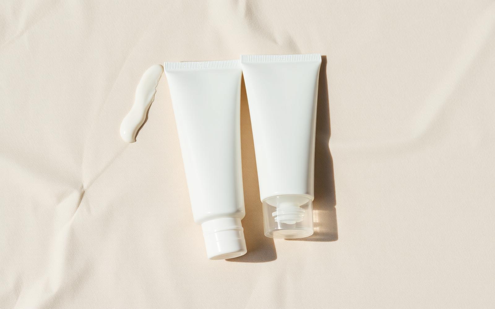 Two plain white sunscreen tubes side by side on pale sand linen with a thick cream swatch beside them