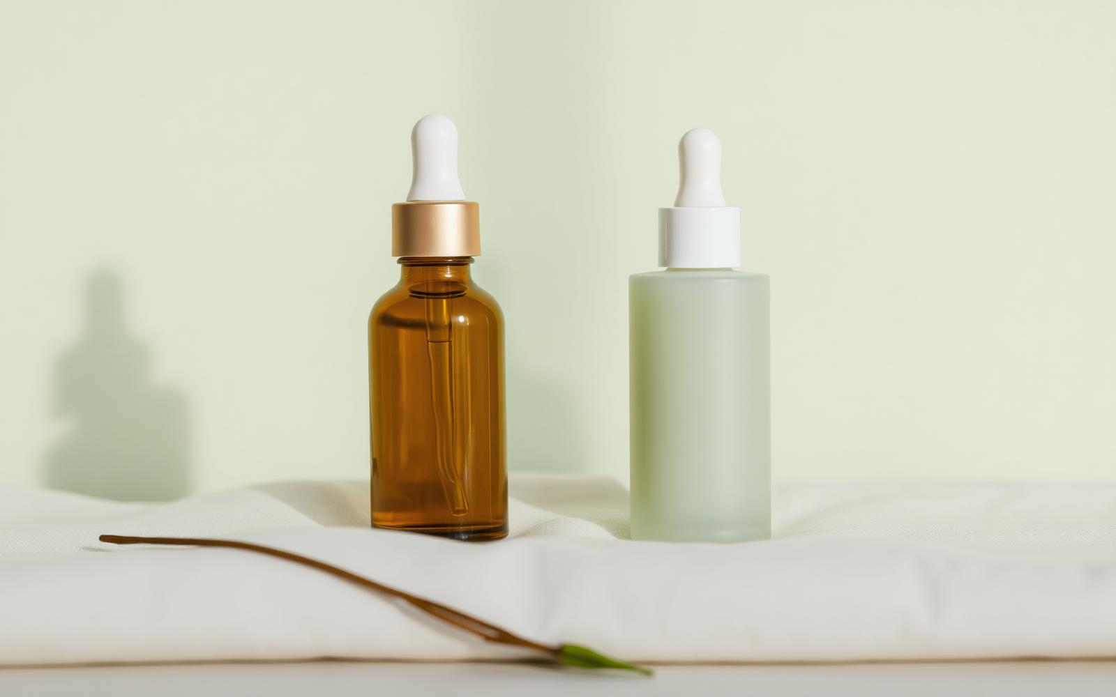 An amber glass dropper bottle beside a pale green frosted serum bottle on cream linen against a soft sage backdrop